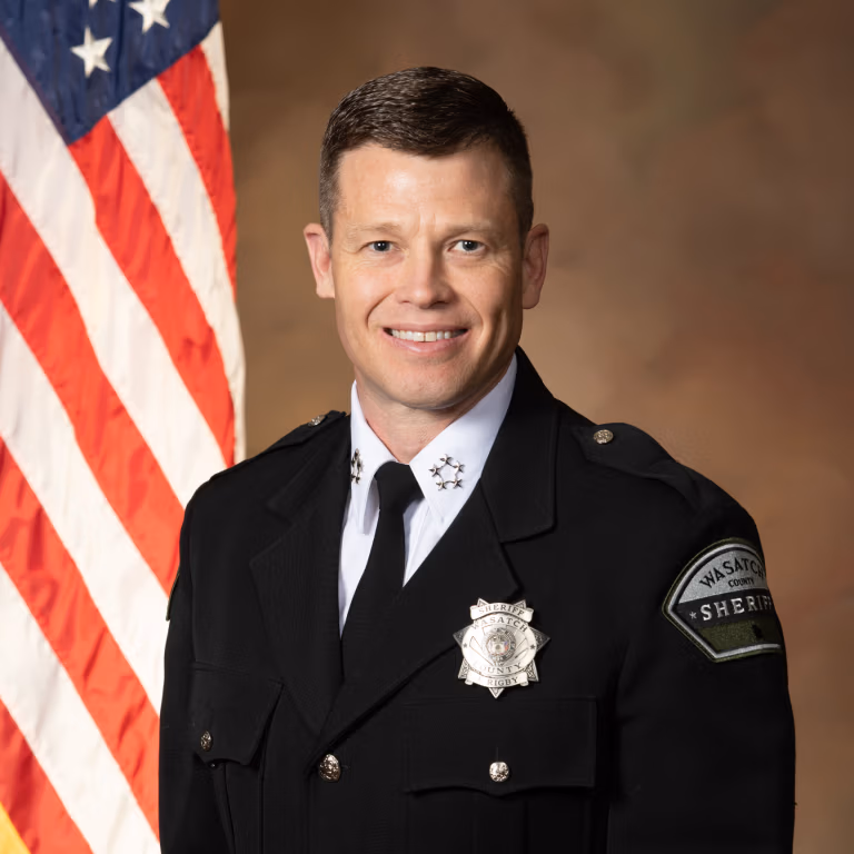 Jared Rigby, wearing a sheriff's uniform with badge, smiling confidently in front of an American flag, representing his campaign for sheriff in Wasatch County, focusing on community safety and law enforcement improvements.