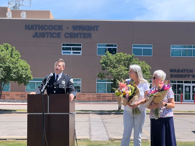 Sheriff speaking at podium in front of Hathcock-Wright Justice Center, two women holding flower bouquets beside him, community event in Wasatch County.