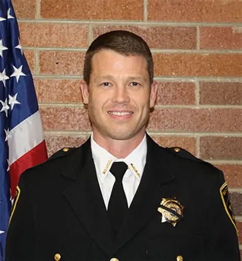 Jared Rigby in sheriff's uniform smiling, standing next to an American flag, representing dedication to public service and community safety in Wasatch County.